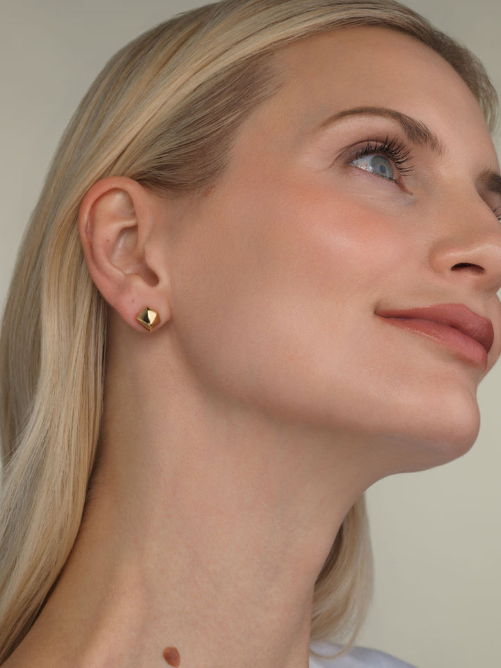 A close-up of a blonde woman wearing a small geometric gold vermeil stud earring by Parkford, on a neutral background.